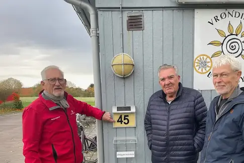 Tre mænd står ved et nummerskilt ved en grå bygning